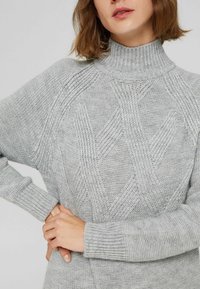 Pull gris clair en maille avec un col montant, présentant des motifs en diamants texturés et des poignets côtelés. Le matériau semble doux et confortable.