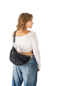 Černá látková kabelka přes tělo s křivkovým tvarem, vybavená přední kapsou a stříbrnými detaily, nošena přes zkrácený bílý texturovaný top a džíny.