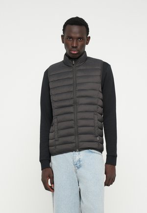 Chaleco de plumas negro acolchado con cuello alto y cremallera frontal, que se lleva sobre una camiseta negra de manga larga. Pantalones de mezclilla azul claro.