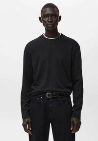 Črna majica z dolgimi rokavi in okroglim izrezom, izdelana iz gladkega blaga. Nošena s temnimi kavbojkami in črnim pasom, z nežnimi detajli šivov.