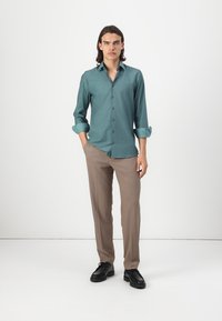 Türkisenes Button-Down-Hemd mit hochgekrempelten Ärmeln, kombiniert mit hellbraunen Hosen und schwarzen Schuhen. Klare Linien und glatte Stoffstruktur.
