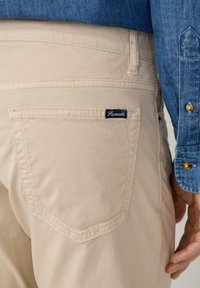 Beige katoenen broek met een achterzak voorzien van een blauwe label. De stof is soepel met subtiele stiksel details. Het shirt is denim blauw.