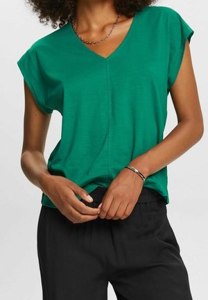 Person med krøllet hår iført en grøn V-hals kortærmet top og sorte bukser, hænderne foldet ved livet med sølvarmbånd og kædehalskæde.