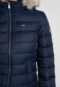 Tmavě modrá péřová bunda s kapucí lemovanou umělou kožešinou, zapínáním na zip a logy. Materiál je lesklý s prošívanou texturou a postranními kapsami.