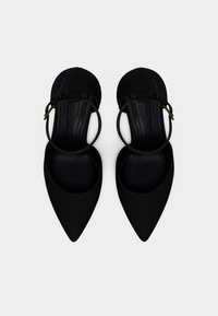 Ζευγάρι μαύρα μυτερά φλατ παπούτσια με λουράκια στον αστράγαλο και μικρή χρυσή αγκράφα σε λευκό φόντο.