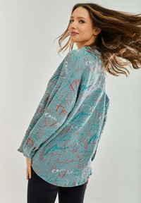 Long-sleeve shirt in textured teal fabric, featuring multicolored script patterns in red, blue, and white. Loose fit with a curved hem.
