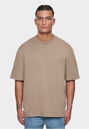 Beige oversized T-shirt van katoen, met een ronde hals, korte mouwen en subtiele branding aan de voorkant, gecombineerd met lichtblauwe denim jeans.