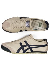 Beige leather sneakers with navy accents, featuring a lace-up design, suede toe cap, and a textured insole with a circular pattern.