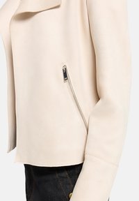 Beige suède cropped jas met een reverskraag, een zilveren ritszakdetail aan de zijkant en een gladde textuur. Zwaar ontwerp.