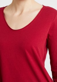 Maglietta rossa a maniche lunghe realizzata in morbido cotone con scollatura a V. Presenta una trama liscia e una minima rifinitura delle cuciture.