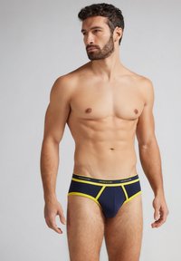 Marineblaue Unterhose mit gelben Akzenten und elastischem Bund, der das Markenlogo trägt. Glattes Material, enganliegendes Design und minimalistischer Stil.