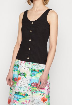 Nainen, jolla on yllään musta hihaton ribbikuvioinen toppi napilla, yhdistettynä värikkääseen hameeseen, jossa on kukka- ja maisemakuoseja.