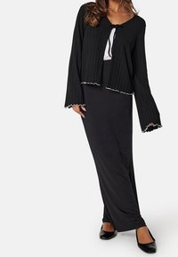 Cardigan nero a coste con maniche a campana e chiusura a fiocco, abbinato a una lunga gonna nera. La texture è liscia e l'orlo ha un bordo arricciato.