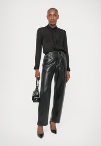 Schwarzes, transparentes Knopfhemd mit vertikalen Streifen, kombiniert mit hoch sitzenden, glänzenden schwarzen Lederhosen und spitzen Absätzen. Kleine schwarze Handtasche.