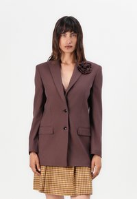 Hnědý blazer se strukturovaným designem, s jedním knoflíkem, dvěma předními kapsami a 3D růží na levém klopě. Nosený přes kostkovanou sukni.