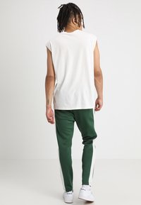 Vit ärmlös skjorta i kombination med gröna joggers med vita sidosträck och vita sneakers, sedd bakifrån.