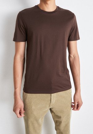 Uomo con maglietta marrone a tinta unita a maniche corte e pantaloni beige di velluto a coste, in piedi su uno sfondo chiaro neutro.