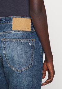 Dżinsy z ciemnoniebieskiego denimu, z jasnobrązową skórzaną naszywką z napisem "rag & bone" oraz kontrastowym żółtym przeszyciem, z widocznymi tylnymi kieszeniami.