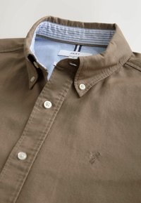 Bruine overhemdbloes met een gestructureerde kraag, lichtblauwe gestreepte binnenvoering en witte knopen. Heeft een klein geborduurd detail.