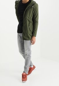 Zielona lekka kurtka z kapturem i kieszeniami na zamek, noszona na czarnej koszuli, w połączeniu z jasnoszarymi jeansami i czerwonymi trampkami.