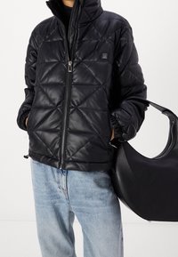 Czarna pikowana kurtka puchowa z błyszczącego materiału z wysokim kołnierzem i zapięciem na zamek, w połączeniu z jasnoniebieskimi dżinsami. W zestawie czarna torebka.