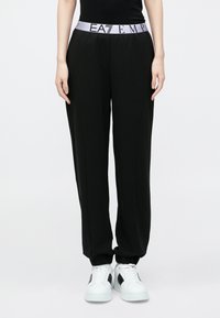 Čierne tepláky so širokým fialovým pásom s nápisom „EAT EMP“ v čiernej farbe. Mäkká, pružná látka so zúženými členkami, kombinované s bielymi teniskami.