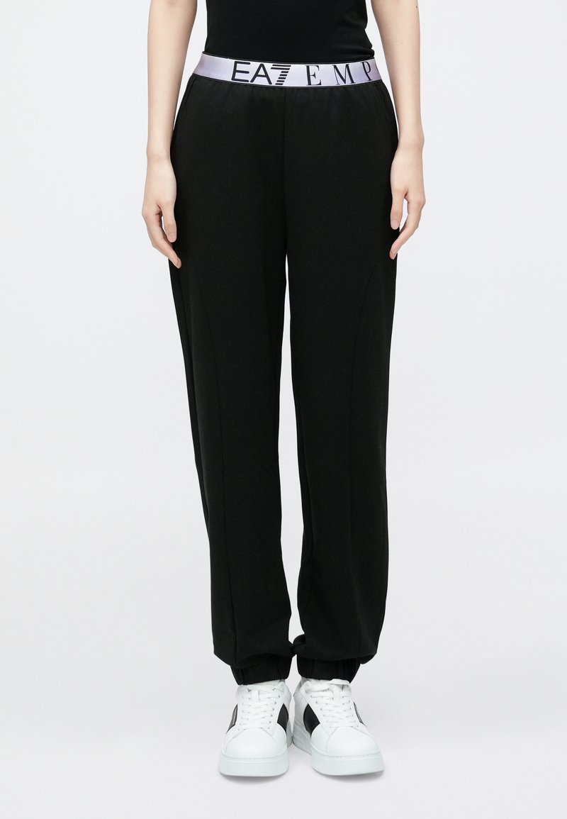 Čierne tepláky so širokým fialovým pásom s nápisom „EAT EMP“ v čiernej farbe. Mäkká, pružná látka so zúženými členkami, kombinované s bielymi teniskami.