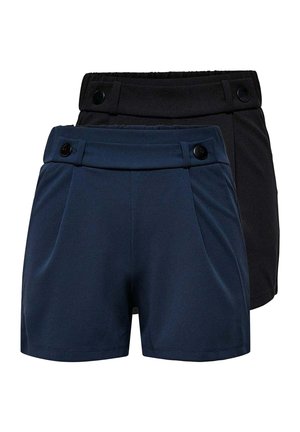 Junge Frau trägt ein weißes Tanktop und marineblaue Shorts, hält eine große schwarze Handtasche, steht und lächelt vor einfachem Hintergrund.