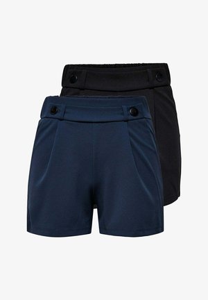 Junge Frau trägt ein weißes Tanktop und marineblaue Shorts, hält eine große schwarze Handtasche, steht und lächelt vor einfachem Hintergrund.