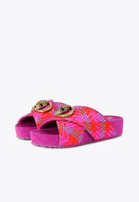 Woven slide sandals feature a pink and red pattern, suede texture, and gold circular hardware embellishments on the straps.