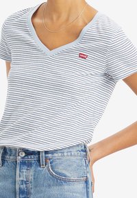 Kék és fehér csíkos V nyakú póló rövid ujjakkal. Bal mellrészen egy kis piros Levi's logó található. Magas derekú farmernadrággal párosítva.