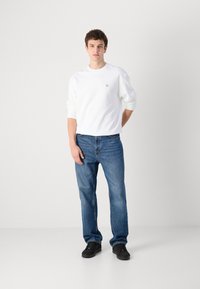 Sudadera blanca de cuello redondo, jeans de mezclilla azules, zapatos negros. Diseño simple con un pequeño logo en el pecho. Ajuste informal, ligeramente holgado.