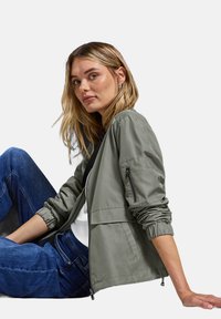 Giovane donna con capelli biondi in giacca verde con zip e jeans blu, seduta di lato, appoggiando una mano contro uno sfondo neutro.
