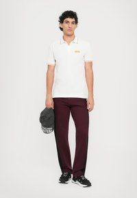 Chemise polo blanche avec des accents jaunes, associée à un pantalon bordeaux. Le modèle tient une casquette gris foncé avec un logo. Des chaussures de sport noires complètent le look.