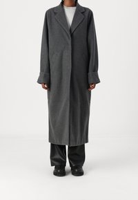 Lungo cappotto grigio in lana con colletto a revers, tasche laterali e ampi polsini. Indossato su un top grigio chiaro e pantaloni neri, dal design minimalista.
