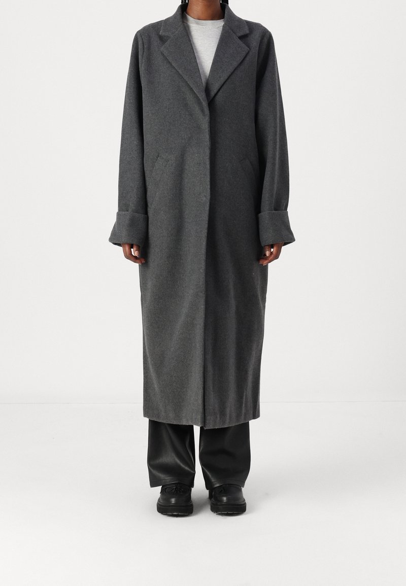 Lungo cappotto grigio in lana con colletto a revers, tasche laterali e ampi polsini. Indossato su un top grigio chiaro e pantaloni neri, dal design minimalista.