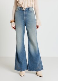 Weit geschnittene blaue Jeans aus Denim, mit einem hellen Waschlook und aufwändiger Stickerei in der Nähe des Saums, kombiniert mit einem cremefarbenen Oberteil und nudefarbenen High Heels.