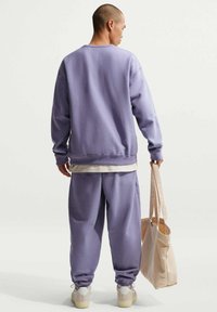 Sweat-shirt violet clair et pantalon de survêtement assorti avec poignets élastiques. Sac fourre-tout beige à la main. Tissu lisse, coupe décontractée, design minimaliste.