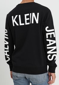 Person wearing black sweatshirt with white "KLEIN" on back, "CALVIN" on left sleeve, and "JEANS" on right sleeve, paired with blue jeans.