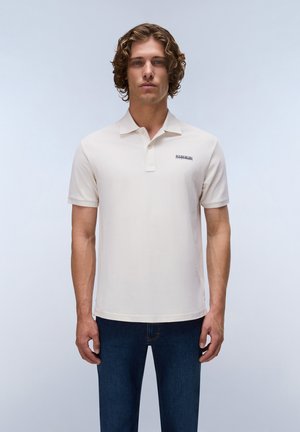 Jeune homme aux cheveux bouclés, portant un polo uni beige clair et un jean bleu foncé, sur un fond clair et uni.