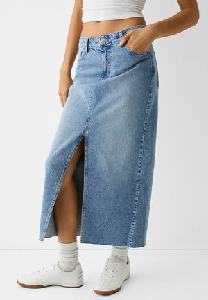 Kvinde iført en lyseblå denimnederdel med høj talje og frontslids, hvid cropped top, hvide sokker og hvide sneakers.
