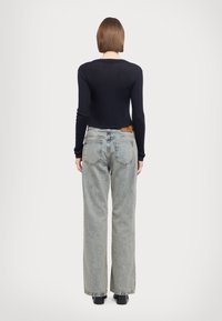 Osoba z prostymi brązowymi włosami, ubrana w czarną prążkowaną bluzkę z długim rękawem, jasnoniebieskie szerokie dżinsy i czarne buty, stojąca plecami.