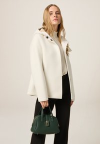 Giacca bianca in misto lana con cappuccio, caratterizzata da bottoni dorati e un accento floreale. Tieni in mano una borsa rigida verde scuro con manici superiori.