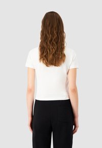 Valkoinen lyhythihainen cropped T-paita, valmistettu pehmeästä kankaasta. Muotoiltu pyöreällä kaula-aukolla, yhdistetty mustiin housuihin, joissa on takatasku.