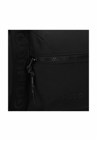 Sort nylon taske med en lynlås lomme, præget logo og en struktureret rem med "GO HUGO" branding. Glat overflade, slankt design.