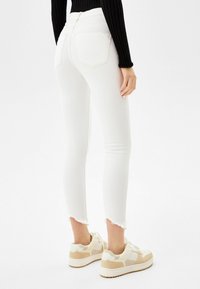 Tejanos ajustados de talle alto en blanco con dobladillo sin terminar, bolsillos traseros y una textura suave, combinados con zapatillas de color claro.