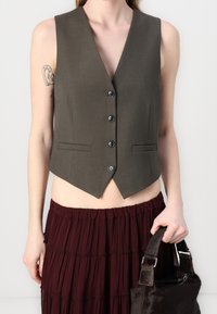 Ciemnooliwkowa bezrękawnik z głębokim dekoltem w serek, pięcioma guzikami i dwiema przednimi kieszeniami. Połączony z bordową, warstwową spódnicą i ciemną torbą.