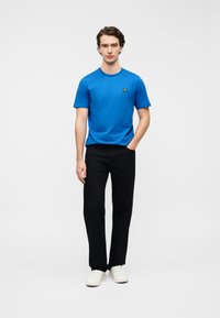 Μπλε βαμβακερό T-shirt με στρογγυλή λαιμόκοψη και μικρό κίτρινο λογότυπο στο στήθος, συνδυασμένο με μαύρο παντελόνι και λευκά αθλητικά παπούτσια.