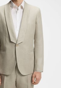 Homme portant une veste de costume beige clair avec un col châle sur une chemise blanche, montré des épaules jusqu'à mi-cuisse.