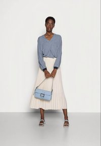 Blusa azul com padrão, saia plissada creme, bolsa azul com fecho quadrado e sandálias com tiras a condizer. Fundo neutro.